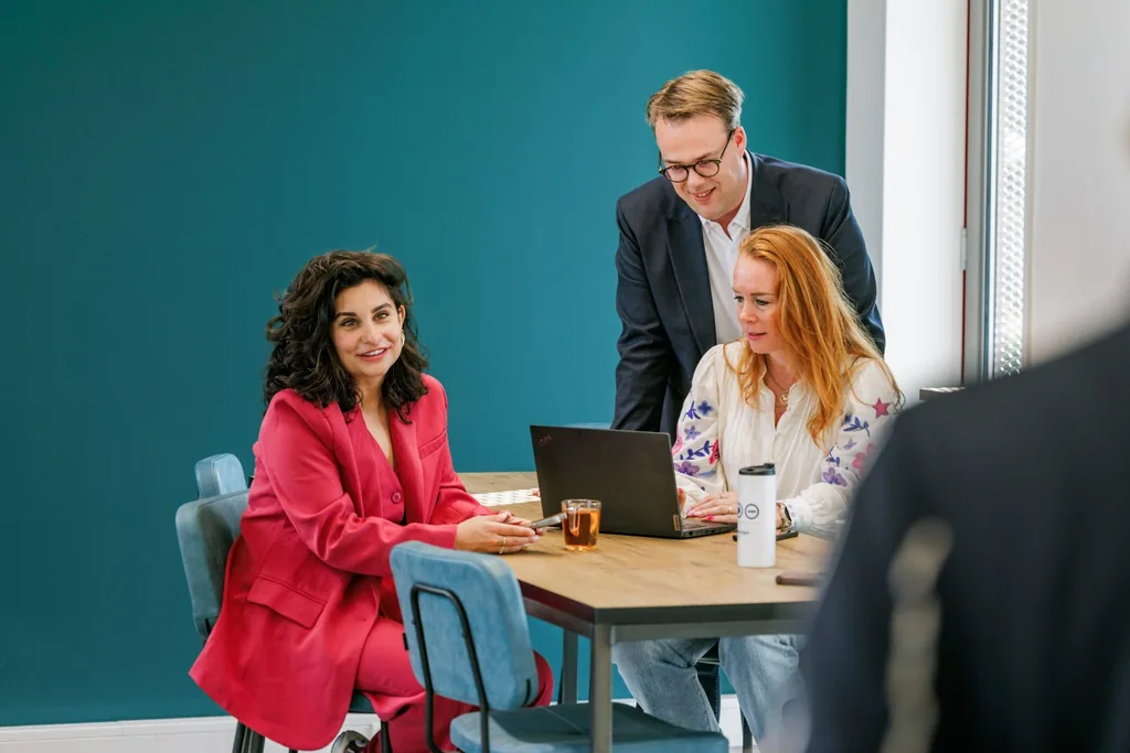 Een collega kijkt lachend in de camera terwijl twee collega’s samen naar een laptop kijken.